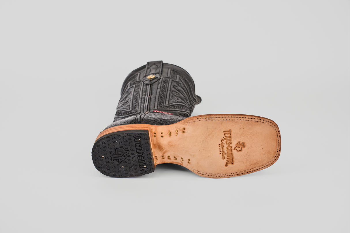 The Exotic Caiman Belly - Black Square Toe boot lies on its side, highlighting the detailed upper stitching, textured black rubber heel, and smooth leather sole featuring branded logos.