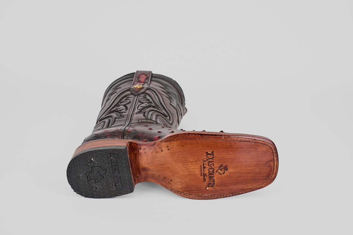 The Exotic Ostrich - Black Cherry - Rodeo Toe boot lies on its side with a Texas-shaped tread and Texas Champion logo visible on the leather sole, showcasing intricate stitching.