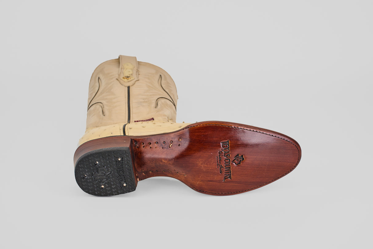 The Exotic Full Quilt Ostrich - Bone - Round Toe boot lies on its side, displaying decorative stitching, a textured genuine ostrich leather pattern, brown sole with “LUCCHESE SINCE 1883” logo, and a black heel with visible nails.