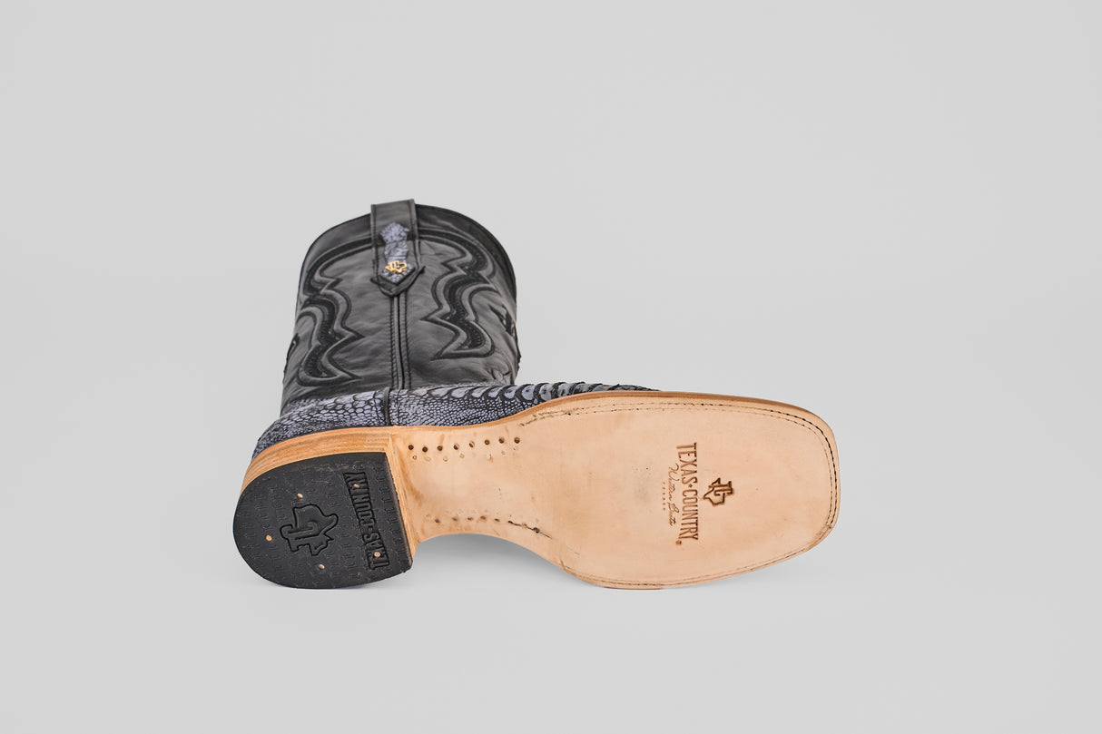 The Austin Leg - Blue Jean - Square Toe men's cowboy boot rests on its side, revealing a light tan leather sole with the Texas Boot Company logo and a black heel, set against a plain light gray background—ideal for blue jeans.