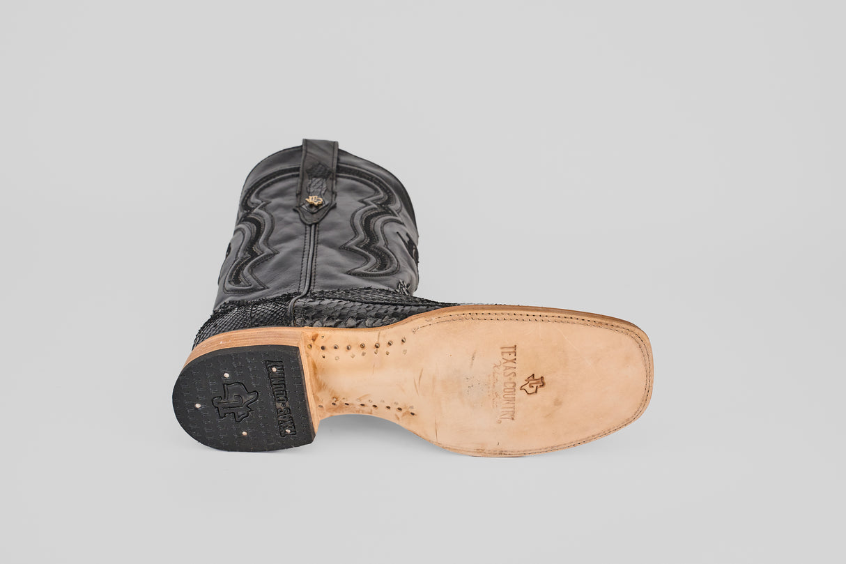 A single Tanner - Black - Square Toe boot lies on its side, showing the light tan sole with "TEXAS SCRIPT" and logo. The patterned black upper and treaded heel are clearly visible against a plain background.