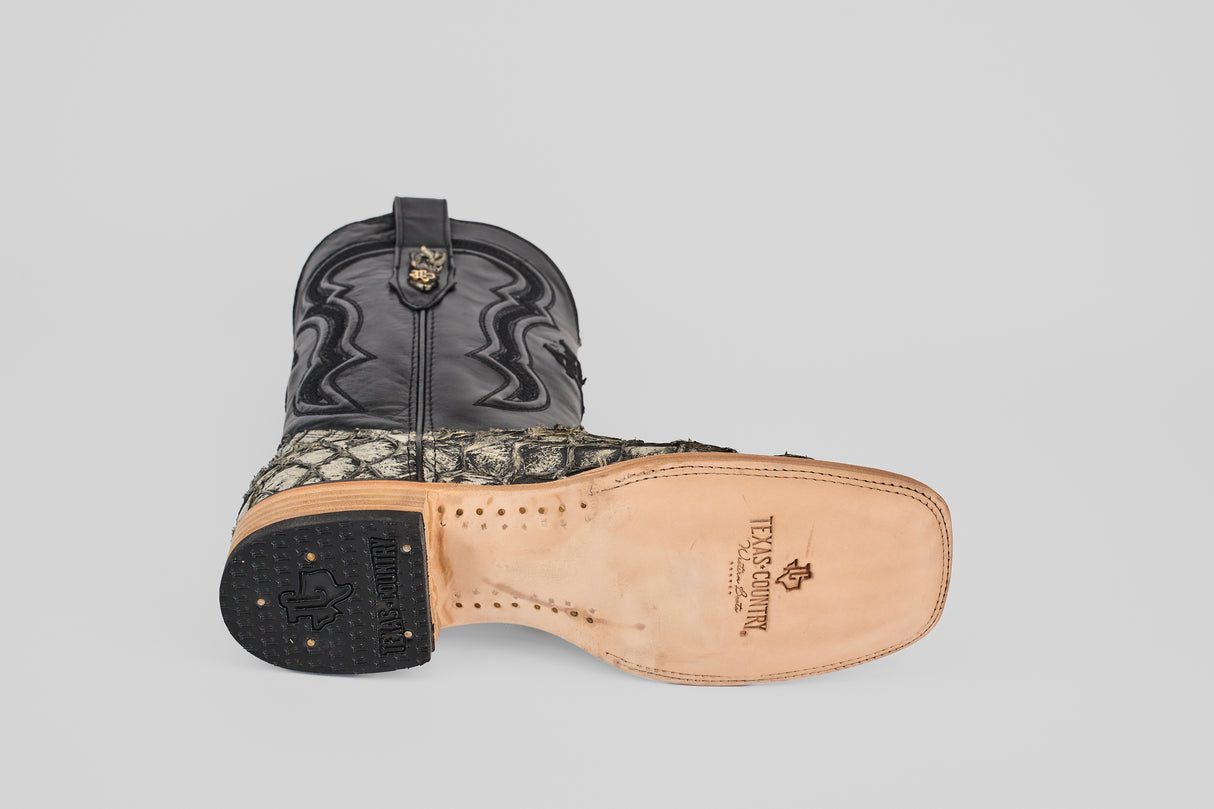 The Conor - Rustic Bone - Square Toe men's cowboy boot lies on its side, displaying the tan leather sole with branding. The black upper features decorative stitching, while the lower showcases a textured fish leather-inspired pattern.