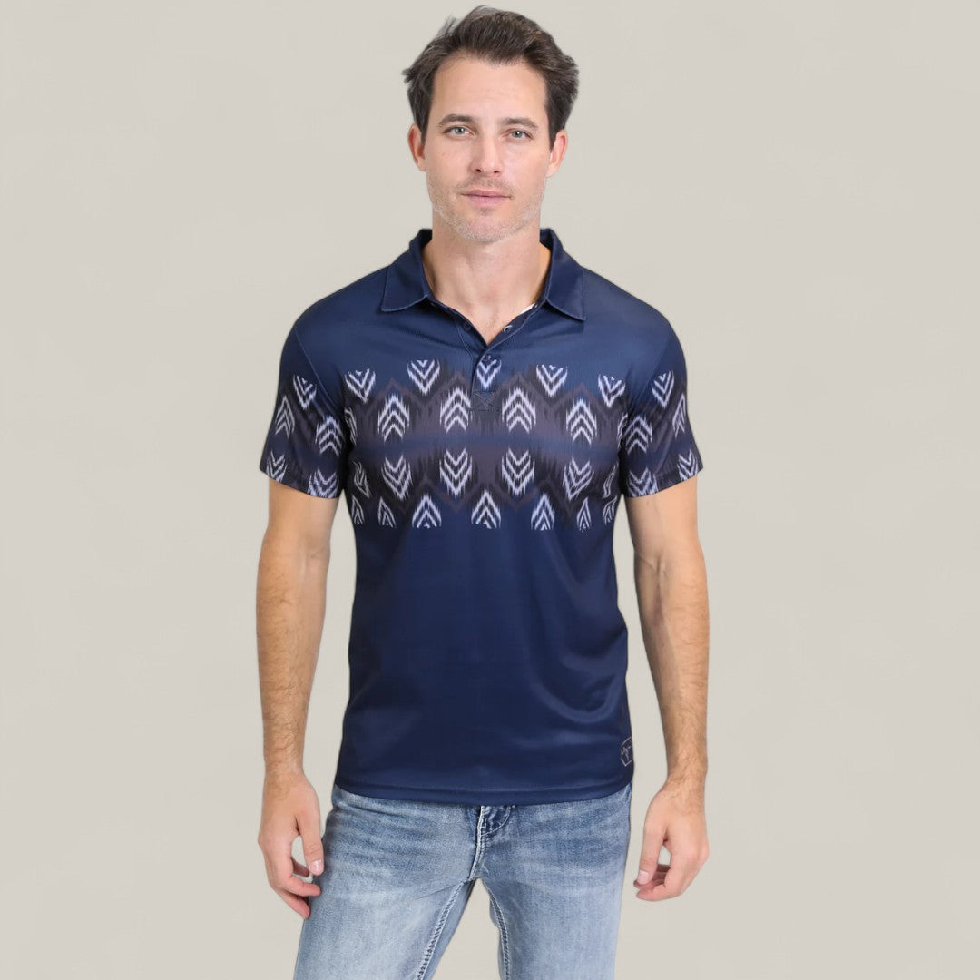 A man wears the Mens Performance Fabric Aztec Panoramic Print Navy Polo (PGP10606) with faded blue jeans, standing against a plain light gray background.