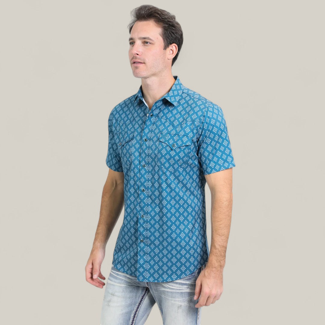 A man wears the Mens Performance Western Short Sleeve Aztec Print Blue Shirt - PGW10632, featuring a white geometric pattern, with light-wash jeans against a plain light gray background.