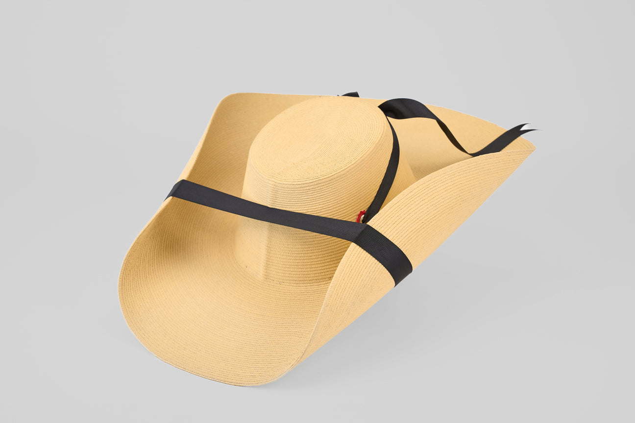A wide-brimmed, light tan straw hat with a black ribbon wrapped around the crown and chin straps, set against a plain light gray background.