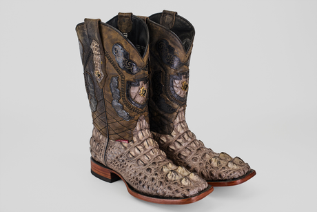A pair of brown crocodile-patterned cowboy boots stand next to a brown leather briefcase on a wooden shelf, against a marble-patterned wall.