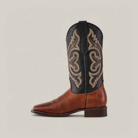 The Grisly Tan - Square Toe boot, made from premium leather, features a profile of a single cowboy boot. Its brown textured lower and decorative black upper with detailed tan stitching highlight its classic Western style against a plain gray background.