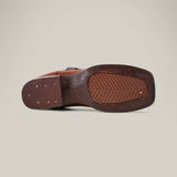 The image features the sole of a Grisly Tan - Square Toe boot, made of premium leather with stitching and small perforations. This Western-style shoe has a textured grip and rectangular heel, set against a plain light gray background.