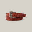 A rolled-up Cognac belt, made with ostrich print and featuring an intricate Texas buckle, set against a plain light gray background.