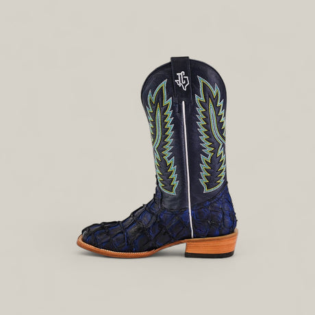 A single Navy Black Exotic Pirarucu Fish - Square Toe boot, from the Platinum Collection, features a textured scale-look lower, green and blue stitching, a white stripe and logo, plus a tan wood heel—luxury boot shown in profile on a neutral background.
