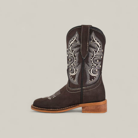 The Amara Suede Oxford - Square Toe - Kids Boots features quality craftsmanship with a dark brown suede finish, intricate white and silver stitching on the shaft, a rounded toe, and a light brown stacked heel against a beige background.