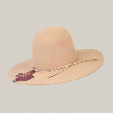 The 10x Belly - TC Hats is a wide-brimmed beige felt Western hat with a simple matching band around its shapeable crown, shown on a light background. A maroon leather tag with gold lettering hangs from the brim.