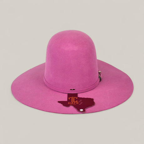 The 4x Pink - TC Hats is a tall-crowned, wide-brimmed pink Western hat with a decorative band and a Texas-shaped TC HATS tag, shown on a light beige background.