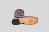 A single Exotic Caiman Horn Back - Black Cherry - Square Toe boot lies on its side, revealing the tan leather sole with the Texas Signature logo, black heel, and decorative stitching on dark leather. The background is plain light gray.