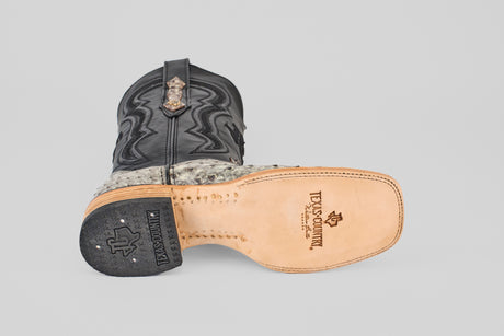 A cowboy boot from Exotic Full Quilt Ostrich - Nicotine - Square Toe lies on its side, showcasing a tan sole with the Texanosmith logo, black leather upper with decorative stitching, and an exotic quilted ostrich pattern near the ankle.