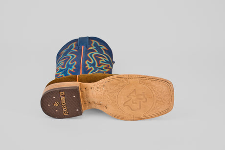 The Gold Suede - Square Toe is a luxury cowboy boot with a tan leather sole featuring TEXAS COUNTRY branding, a decorative emblem, and an upper crafted from blue leather with colorful embroidered patterns.