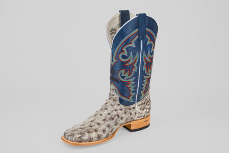 The Russell - Rustic Gray - Square Toe boot features a textured light gray ostrich leather foot, a tall blue shaft with red and white stitching, and a light brown wooden heel, all shown on a plain gray background.