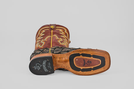 The Butch - Caramel - Square Toe cowboy boot rests on its side, displaying its detailed stitching, leather sole with rubber heel grip, and unique two-tone caramel and black design crafted in rich Pirarucu leather with decorative upper patterns.