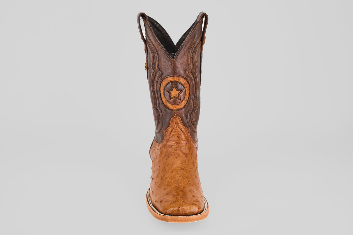 The Exotic Full Quilt Ostrich - Brandy Brucciato, a single square toe cowboy boot in rich brown with a decorative star emblem on the front, is displayed upright against a plain light gray background.