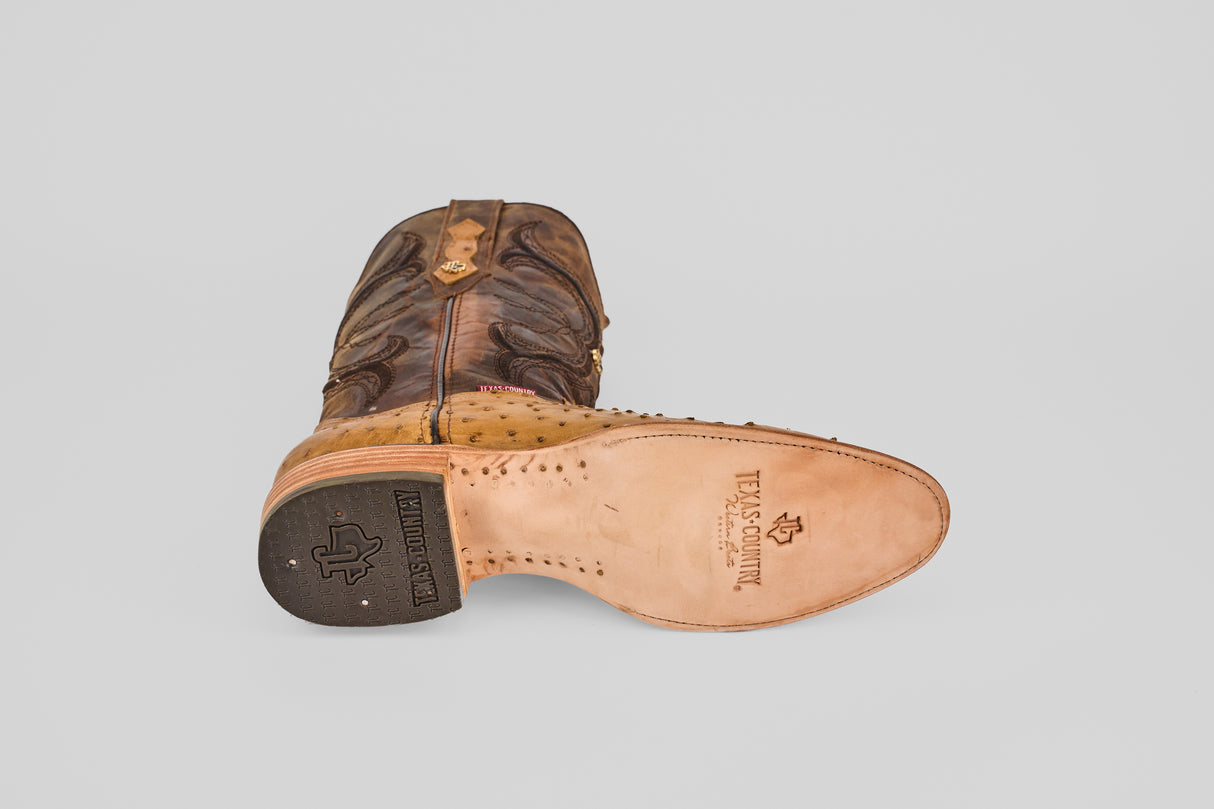 The Austin - Antique - Roper Toe boot, in brown and tan ostrich leather, lies on its side, displaying a sole with "Texas Country" and a Texas map; the heel shows an embossed logo.