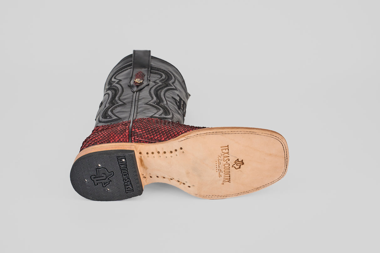 The Tanner – Scarlet – Square Toe cowboy boot lies on its side, revealing a tan sole with TEXAS COUNTRY branding, a black rubber heel, and a textured red and black upper with wavy western stitching on a plain light gray background.