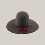 The 10X - Oxford - TC Hats is a wide-brimmed, dark Western hat with a tall, shapeable open crown. It’s displayed on a plain light background and features a red Texas-shaped tag on the brim with small text and graphics.