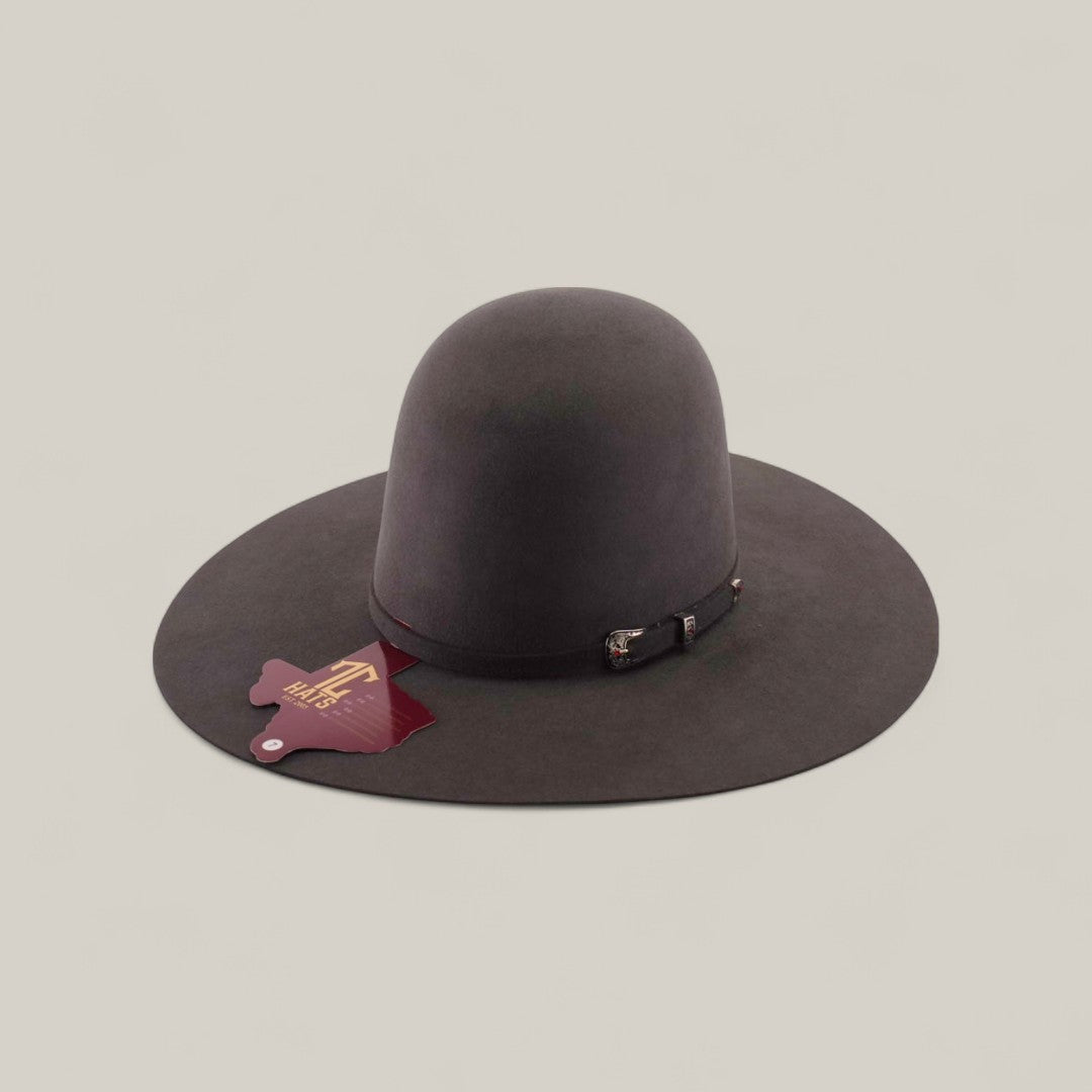 The 10X - Oxford - TC Hats is a wide-brimmed, dark brown Western hat with a shapeable crown and decorative strap, shown against a beige background. A maroon cattle brand-shaped tag hangs from the brim.
