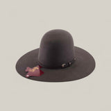 The 10X - Oxford - TC Hats is a wide-brimmed, dark brown Western hat with a shapeable crown and decorative strap, shown against a beige background. A maroon cattle brand-shaped tag hangs from the brim.
