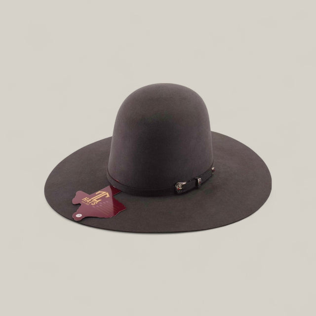 The 10X - Oxford - TC Hats is a wide-brimmed, dark brown Western hat with a shapeable crown and decorative strap, shown against a beige background. A maroon cattle brand-shaped tag hangs from the brim.