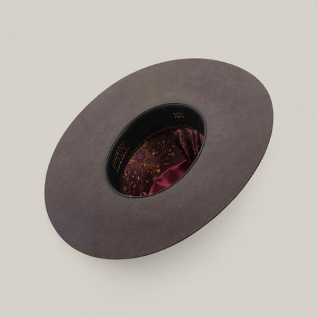 The 10X - Oxford - TC Hat is displayed upside down, featuring a wide gray brim, a black inner band, and a maroon paisley-patterned lining with a shiny accent, set against a plain beige background.