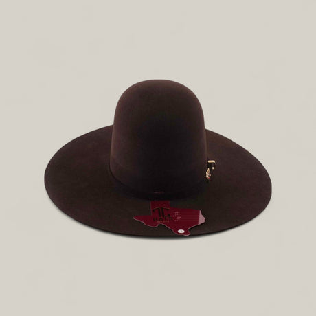 The 10X - Chocolate - TC Hats, a wide-brimmed dark brown Western hat with a decorative band and Texas-shaped tag on the brim, is displayed on a plain, light background.