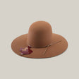 The 4X - Fawn - TC Hats is a light brown felt Western hat with a wide brim, round crown, and thin matching hatband. A maroon Africa-shaped tag hangs from the band. The hat is shown against a plain beige background.