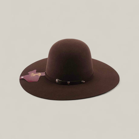 The 4X - Chocolate - TC Hats is a wide-brimmed brown Western felt hat with a rounded crown and decorative band, displayed on a light beige background. A purple cowboy hat-shaped tag is attached to the left brim.