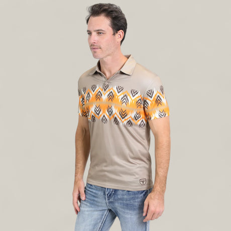 A man stands against a plain light background wearing the Mens Performance Fabric Aztec Panoramic Print Khaki Polo (PGP10607) featuring an orange and black geometric pattern across the chest and sleeves, paired with blue jeans.