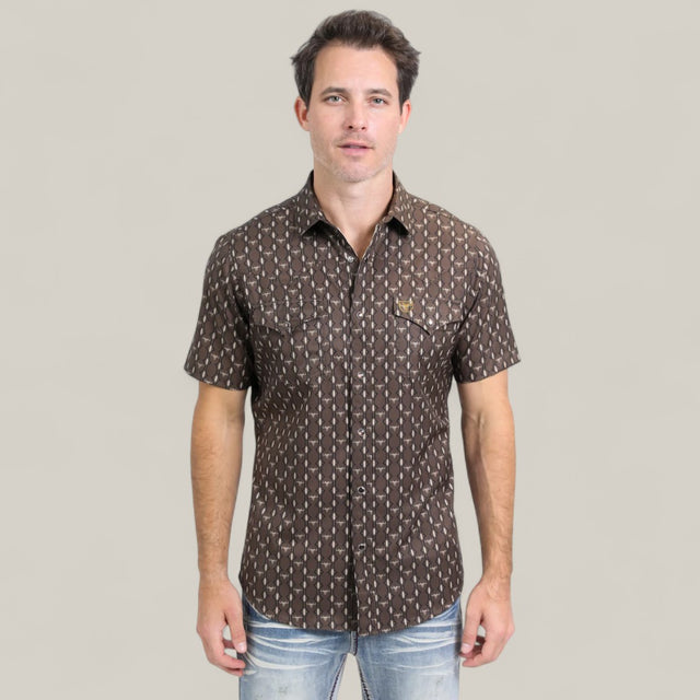 A man wearing the Mens Performance Western Short Sleeve Aztec Print Brown Shirt - PGW10643 and light blue jeans stands against a plain, light gray background, facing forward with a neutral expression.