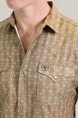 A close-up of a person wearing the Mens Performance Western Short Sleeve Aztec Print Camel Shirt (PGW10645), featuring white geometric designs, two chest pockets (one with an embroidered bull head), and a partially unbuttoned collar.