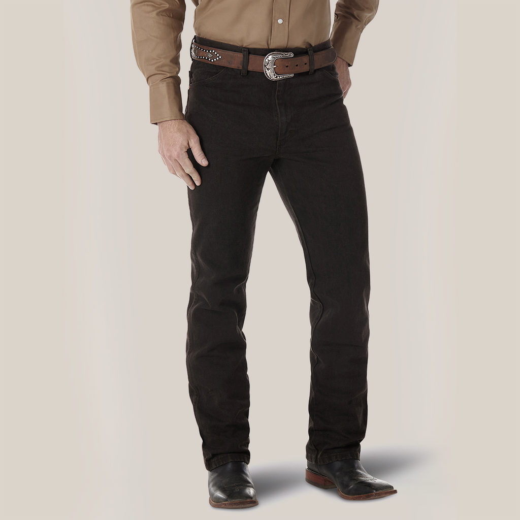 A person wearing a tan long-sleeve shirt, Wrangler Cowboy Cut Slim Fit Jean (0936KCL), a studded brown belt with a large buckle, and black boots. The backdrop is plain white.