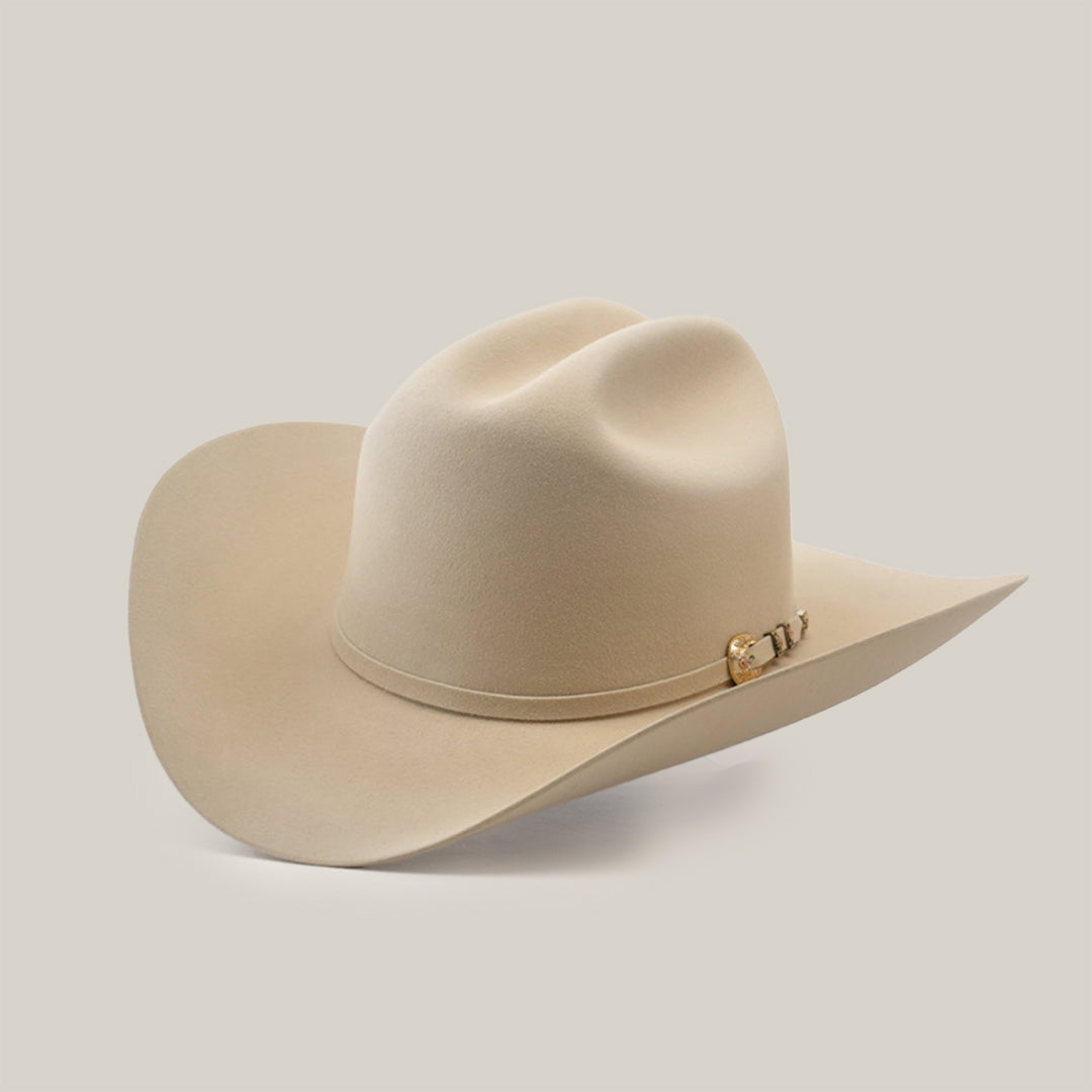 The 500X Superior Belly, a beige cowboy hat made from 100% belly beaver fur, features a wide brim and a decorative band with a metallic embellishment. Displayed on a smooth light gray surface, it is slightly tilted to highlight its premium craftsmanship.