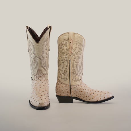 The Karoo Ostrich Print Bone boots, in a light color, feature intricate stitching and an ostrich print texture. One boot stands upright while the other lies on its side to showcase its distinctive heel and side design.