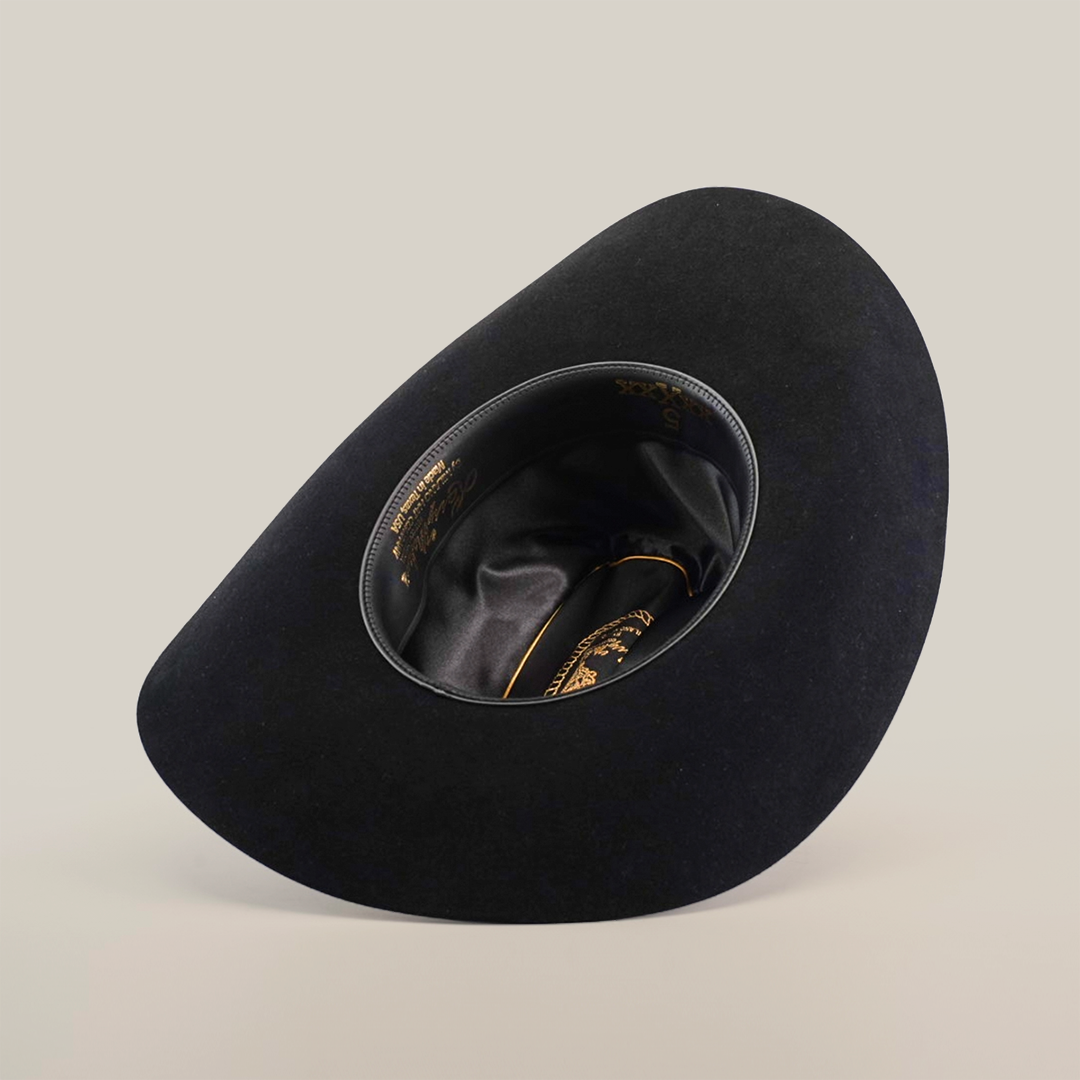 The 5X Dorado Black hat from Milano Hat Co., crafted from wild rabbit and beaver fur felt, rests upside down on a white background. It features a smooth, curved shape with glossy inner lining and gold text near the rim, embodying the elegance of a timeless western headpiece.