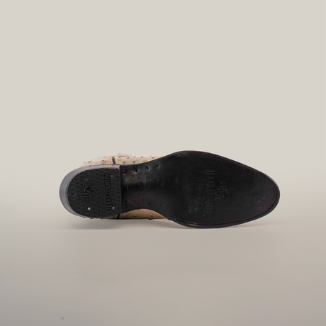 The image features the sole of the Karoo Ostrich Print Bone - Round Toe boot, showcasing a beige handcrafted design with a black bottom. Star-shaped treads decorate the leather sole, with the brand name near the heel. A plain white background highlights its stylish aesthetic.