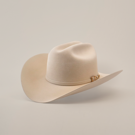 The 100X Independencia Belly, a luxury beige cowboy hat made from premium full beaver fur, boasts a curved brim and decorative band, elegantly showcased on a white surface.